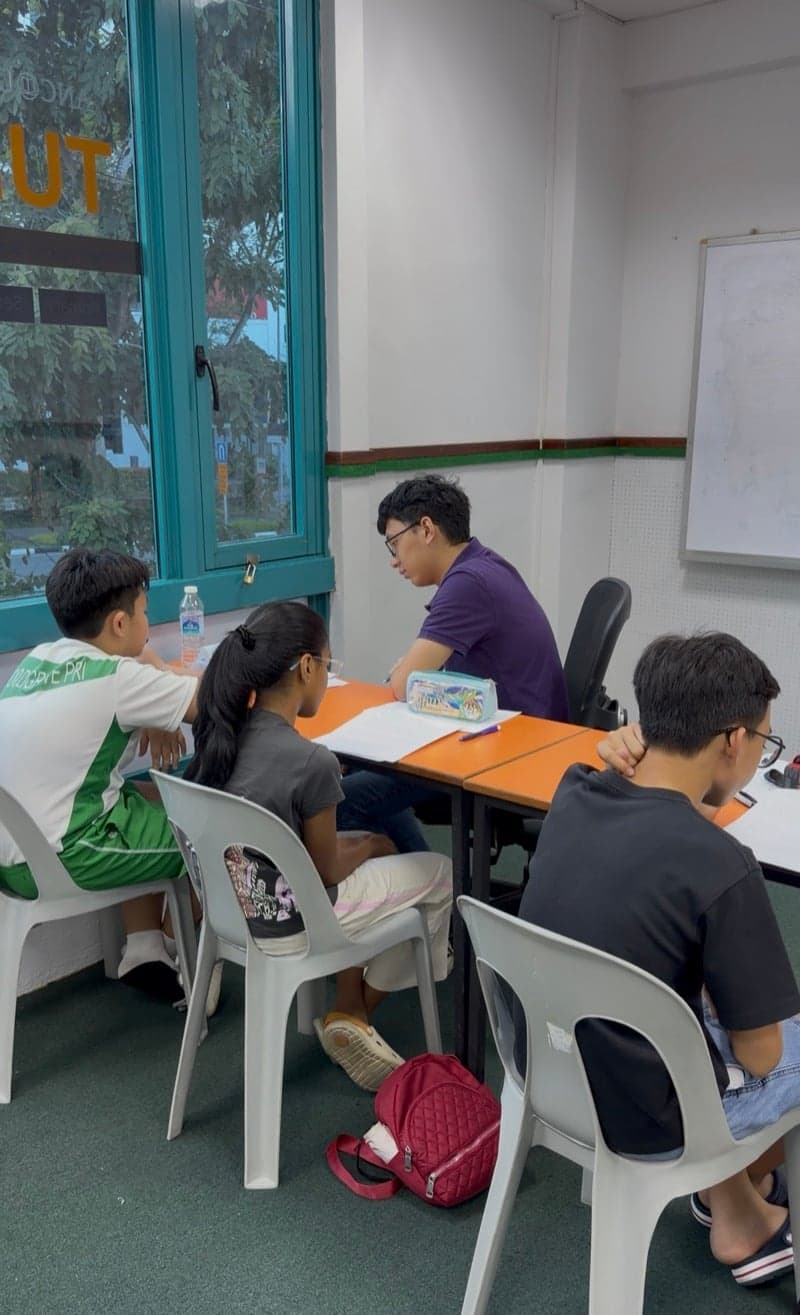 Primary students in a small-group lesson at Ancourage Academy Woodlands, working through a worksheet with their tutor by the teal-framed windows above Woodlands Drive 44.