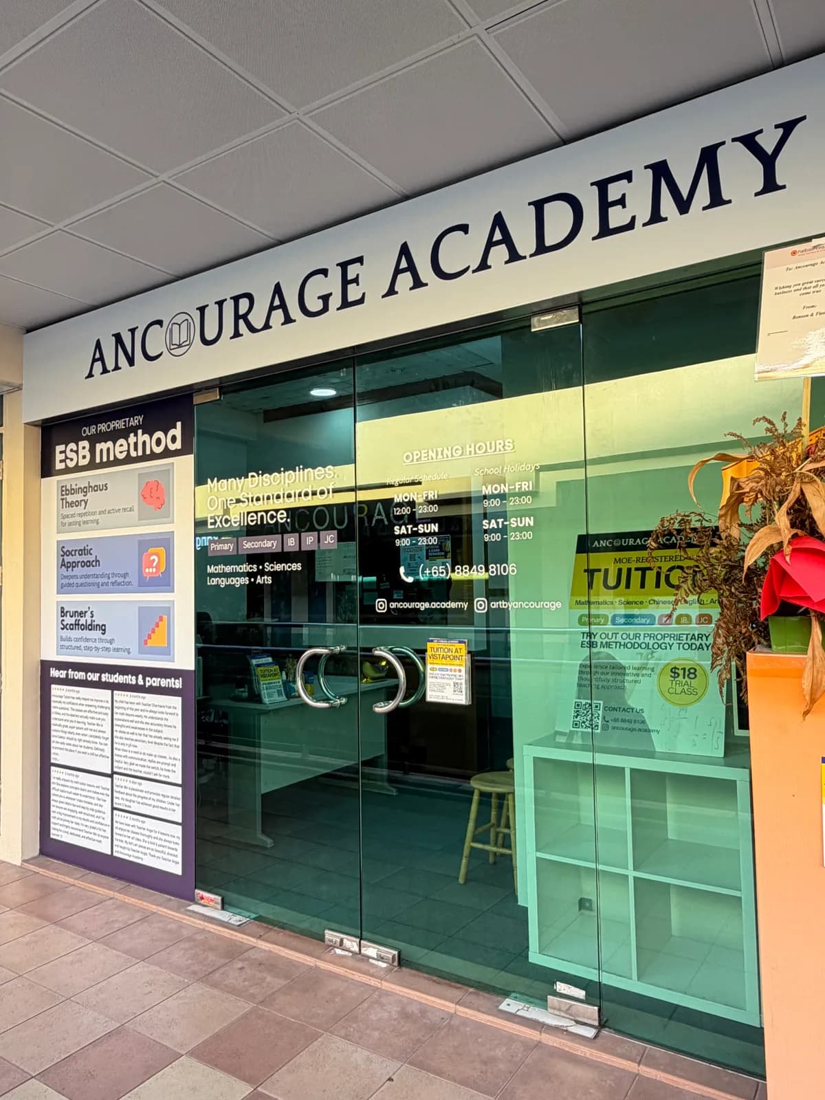 Glass entrance of Ancourage Academy Woodlands showing the opening hours decal — Mon–Fri 12pm–11pm, Sat–Sun 9am–11pm — with phone and Instagram contacts on the door.